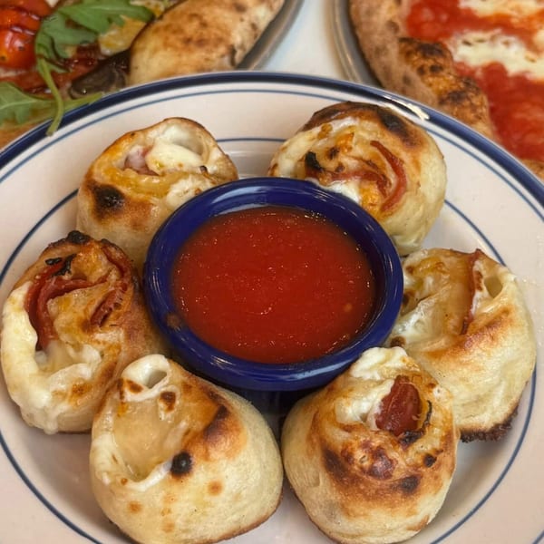 Plate of mini pizza bites surrounding a bowl of tomato dipping sauce, with larger pizzas visible in the background.