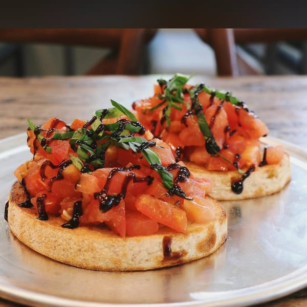 Two toasted bruschetta slices topped with diced tomatoes, fresh basil, and balsamic glaze on a metal plate.